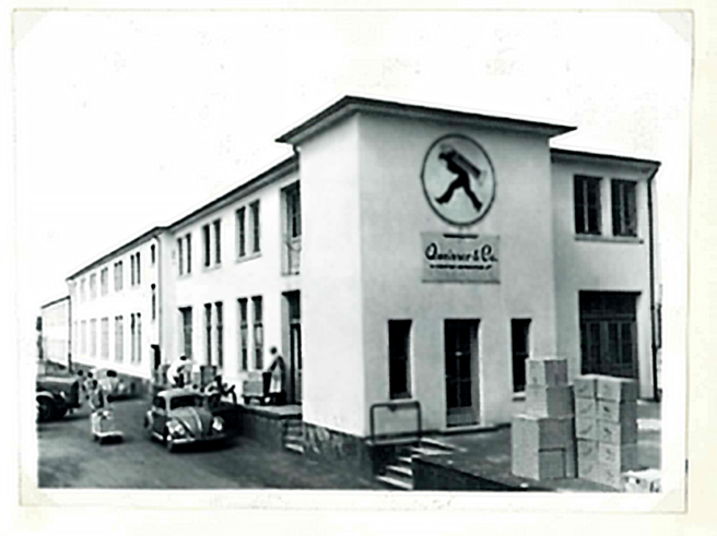 Ein historisches schwarz-weiß Foto eines Gebäudes mit einem großen Schild, das eine stilisierte Figur zeigt. Vor dem Gebäude stehen einige Personen und Autos, während an der Seite Kisten gestapelt sind. Das Gebäude hat große Fenster und eine Eingangstür.