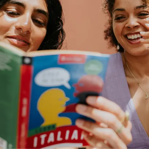 Due giovani donne sorridono mentre leggono un libro di italiano. Una di loro tiene il libro vicino al viso, mostrando la copertina colorata, mentre l'altra guarda con interesse. Sullo sfondo, un muro di colore caldo. L'atmosfera è di gioia e condivisione.