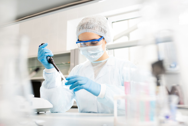 Eine Laborantin in Schutzkleidung, mit Handschuhen und einer Schutzmaske, verwendet eine Pipette, um eine Flüssigkeit in einem Labor zu transportieren. Um sie herum sind verschiedene Laborausrüstungen sichtbar.