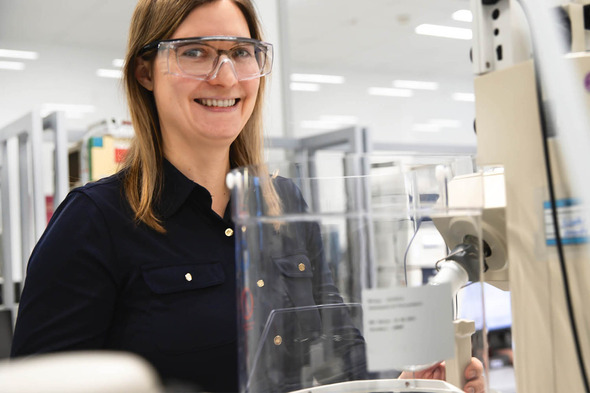 Eine lächelnde Frau trägt Schutzbrille und steht neben einem Laborgerät. Im Hintergrund sind Regale mit wissenschaftlichen Materialien sichtbar. Die Atmosphäre wirkt freundlich und professionell.